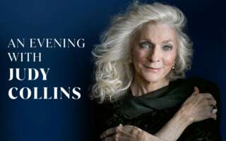 Judy Collins smiles warmly with arms folded across shoulders in front of solid navy blue background